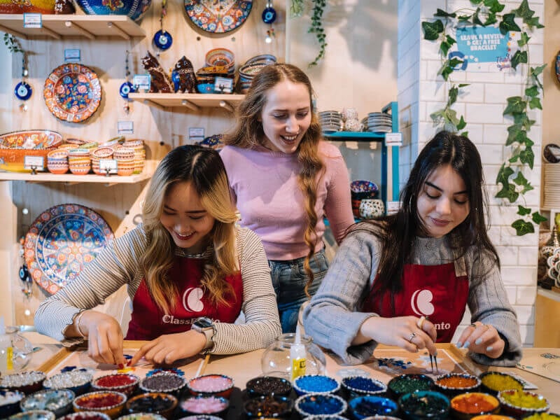 Two seated women making mosaics with teacher behind them